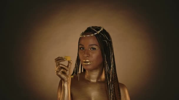 非洲裔美国女人 穿着克利奥帕特拉风格的棕色背景 有圆形的光 一个头戴珠宝 皮肤金黄的女人 吃着一个金黄色的巧克力糖果 时尚艺术设计 慢动作 — 图库视频影像