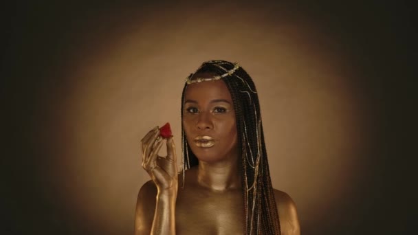 非洲裔美国妇女在工作室里的克利奥帕特拉风格的棕色背景与圆形的光 一个头上戴着金色皮肤和珠宝的女人吃着多汁的成熟草莓 时尚艺术设计 慢一点 — 图库视频影像
