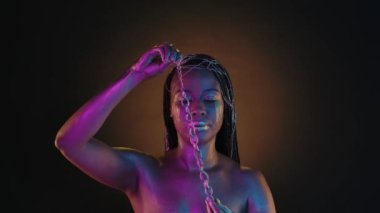 Pembe neon ışıklı Afrikalı bir kadın avucundan altın bir zincir alıp boynuna takıyor. Altın makyajlı bir kadın. Yüzünde altın gözyaşları var. Stüdyoda.