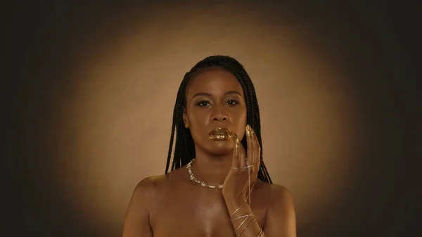 African American woman applies golden paint on her lips, liquid gold. Seminude woman with gold chain bracelets on her hands, with a necklace around her neck on a brown background with circular light