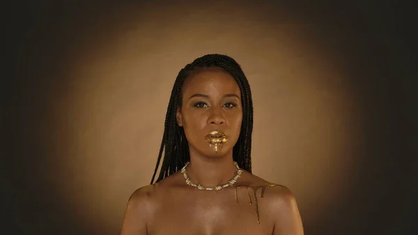 Portrait of an African American woman with drops of liquid gold dripping from her lips and from her bare shoulder. Seminude woman with a golden necklace around her neck on a brown background with