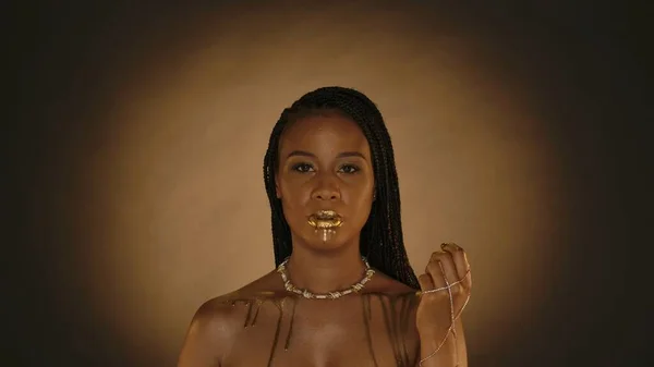 Portrait of a woman with liquid gold dripping from her lips and bare shoulders. A seminaked woman with a golden necklace around her neck, with a golden chain on her hand, fingertips and nails in gold