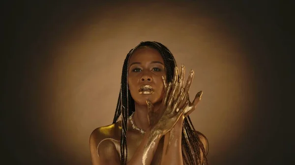 A woman with golden lips, golden skin and golden chains in her hair in the studio on a brown background with circular light. Arms of a seminude African American woman in liquid gold, or gold paint