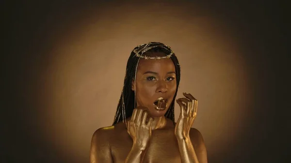 African American woman in Cleopatra style on a brown background with circular light. A woman with golden skin and jewelry on her head holds a golden chocolate candy with her lips, in her mouth
