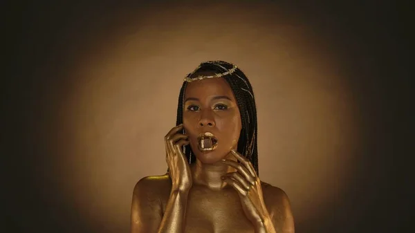 非洲裔美国妇女在克利奥帕特拉风格的棕色背景与圆形光 一个头上戴着金色皮肤和珠宝的女人嘴里衔着一颗含金巧克力糖 — 图库照片