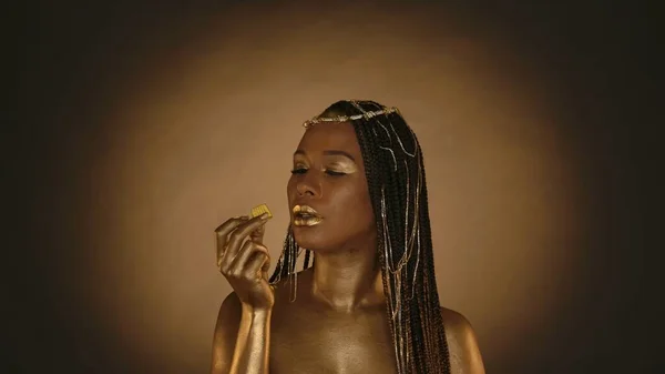 Portrait of an African American woman in Cleopatra style on a brown background with circular light. A woman with golden skin and jewelry on her head holds a golden chocolate candy. Fashion art design