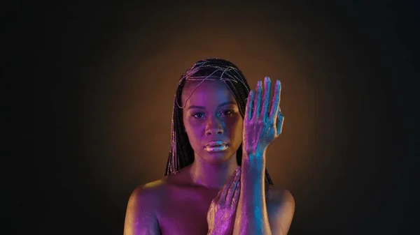 一位非洲裔美国妇女在褐色的背景上用圆形的光展示她的手绘着金色的颜料 在粉色霓虹灯下 拥有富有创意的克利奥帕特拉风格化妆品和金色珠宝的女人 — 图库照片