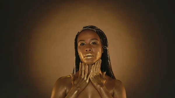 アフリカ系アメリカ人の女性が 金色の塗料に塗られた手で首に触れる 目を閉じたアフリカ系アメリカ人女性が 金色の塗料に塗られた手で顔に触れる — ストック写真