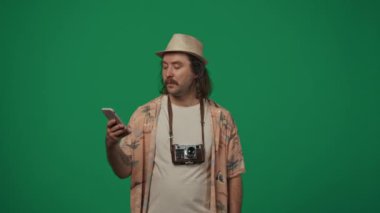 Gezici reklam yaratıcı konsepti. Günlük kıyafetler giymiş, hasır şapkalı, eski kameralı, akıllı telefondan selfie çeken, telefondaki fotoğrafları kontrol eden bir adam. Yeşil arkaplanda izole.