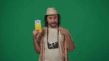 Gezici reklam yaratıcı konsepti. Günlük kıyafetler giyen, şapkalı, elinde uçuş bileti ve pasaport tutan, gülümseyen yüzlü, parmakla işaret eden bir adam. Yeşil arkaplanda izole.