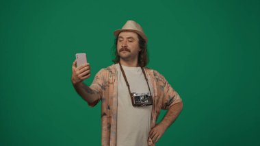 Seyahat eden yaratıcı konsept. Günlük kıyafetlerle gezen, hasır şapkalı, eski kameralı, akıllı telefondan selfie çeken ve gülümseyen bir adam. Yeşil arkaplanda izole.