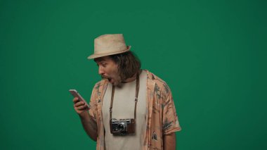 Seyahat eden yaratıcı konsept. Sıradan giyinen ve şapkalı bir adam akıllı telefon tutuyor, ilginç bir şey okuyor, şoke olmuş bir yüz ifadesi. Yeşil arkaplanda izole.