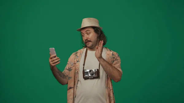 クリエイティブなコンセプトの旅 男はカジュアルな服で旅行し ビデオ通話でスマートフォンで話す帽子をかぶります 緑の背景に孤立した — ストック写真