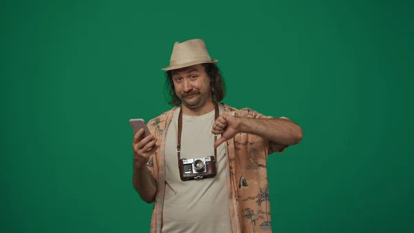 クリエイティブなコンセプトの旅 スマートフォンを保持しているカジュアルな服とわら帽子の男は ネガティブな顔で親指を示しています 緑の背景に孤立した — ストック写真