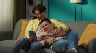 Evde homoseksüel bir çiftin videosu. Kanepede uzanıyorlar, tablet üzerinde fotoğraflar izliyorlar, sarılıyorlar, gülüyorlar ve birbirlerine içtenlikle konuşuyorlar. LGBT, eşitlik, eğitim içeriği, gurur.