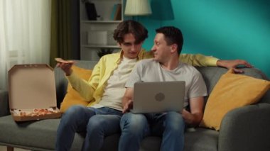 Evde homoseksüel bir çiftin videosu. Kanepede oturuyorlar, dizüstü bilgisayarda fotoğraflar izliyorlar, pizza yiyorlar, sarılıyorlar ve birbirlerine içtenlikle konuşuyorlar. LGBT, eşitlik, eğitim içeriği, gurur.
