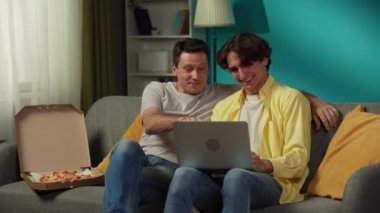 Evde homoseksüel bir çiftin videosu. Kanepede oturuyorlar, dizüstü bilgisayarda fotoğraflar izliyorlar, pizza yiyorlar, sarılıyorlar ve birbirlerine içtenlikle konuşuyorlar. LGBT, eşitlik, eğitim içeriği, gurur.