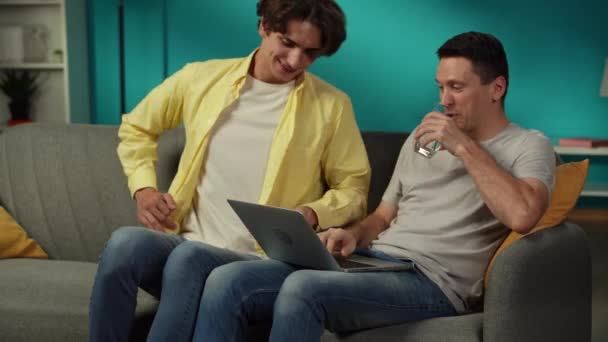 自宅で同性愛者のカップルのビデオ 一人はソファーに座り ラップトップに取り組んでいる 彼のパートナーがフレームに入った 彼らは抱擁し 温かく話しています Lgbt 教育コンテンツ プライド — ストック動画