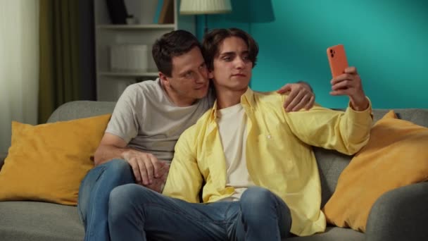2人の同性愛者のビデオ 自宅でカップル 彼らはソファーに座り 抱擁し キスし セルフィーをとって幸せに話し合っています Lgbt 教育コンテンツ プライド — ストック動画