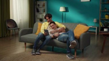 Evde homoseksüel bir çiftin tam boy videosu. Kanepede uzanıyorlar, tablet üzerinde fotoğraflar izliyorlar, sarılıyorlar, gülüyorlar ve birbirlerine içtenlikle konuşuyorlar. LGBT, eşitlik, eğitim, gurur.