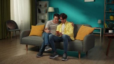 Evde iki homoseksüel adamın tam boy videosu. Kanepede oturuyorlar, sarılıyorlar, selfie çekiyorlar ve mutlu bir şekilde tartışıyorlar. LGBT, eşitlik, eğitim içeriği, gurur.