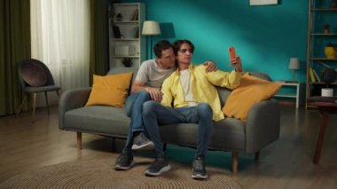 Evde iki homoseksüel adamın tam boy videosu. Kanepede oturuyorlar, sarılıyorlar, selfie çekiyorlar ve mutlu bir şekilde tartışıyorlar. LGBT, eşitlik, eğitim içeriği, gurur.