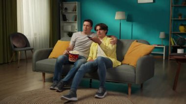 Evde homoseksüel bir çiftin tam boy videosu. Kanepede oturuyorlar, televizyon izliyorlar, ne izleyeceklerini seçiyorlar, patlamış mısır yiyorlar, sarılıyorlar ve birbirleriyle konuşuyorlar. LGBT, eşitlik, eğitim.