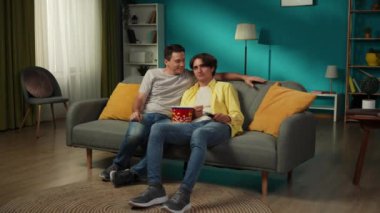 Evde homoseksüel bir çiftin tam boy videosu. Kanepede oturuyorlar, birlikte televizyon izliyorlar, patlamış mısır yiyorlar, sarılıyorlar, gülümsüyorlar, sevgi ve şefkat gösteriyorlar. LGBT, eşitlik, eğitim, gurur