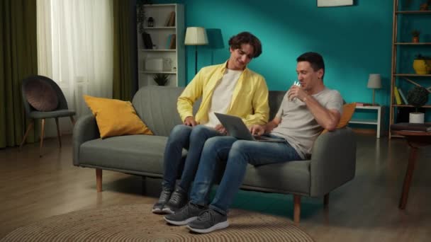 フルサイズ 同性愛カップル 一人はソファーに座り ラップトップに取り組んでいる 彼のパートナーはガラス1杯の水でフレームに入ります 彼らは抱きしめて話している Lgbt プライド — ストック動画