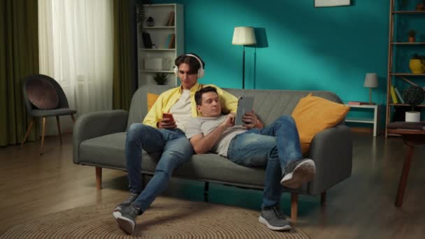 自宅で同性愛カップルのフルサイズのビデオ 彼らはソファーのスクロールに横たわり ネットをサーフィンしています 一つは 彼の好きな曲を共有するために彼のパートナーにヘッドフォンを与えることです Lgbt — ストック動画