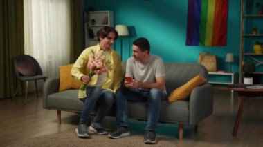 Evde homoseksüel bir çiftin tam boy videosu. Biri elinde çiçek buketi ile çerçeveye girer, bunları partnerine verir, iltifatlar, dileklerle. LGBT, eşitlik, eğitim içeriği.