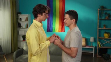 Homoseksüel bir çiftin el ele tutuşup alınlarını birbirine yasladığı orta boy bir video. Arka planda gökkuşağı bayrağı var. LGBT, eşitlik, eğitim, gurur.