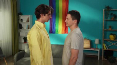Homoseksüel bir çiftin birbirlerinin önünde durup şefkatle sarılıp birbirlerini okşamalarının orta boy videosu. Arka planda gökkuşağı bayrağı var. LGBT, eşitlik, eğitim, gurur.