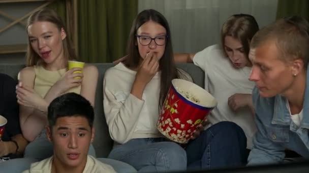 10代 ソファーに座ってテレビを見ている友人 ホラー映画 シリーズ 彼らはポップコーンを食べ チャットし 叫び声に飛び乗っています ティーンエイジ — ストック動画