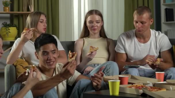 一组青少年 年轻人 坐在沙发上和桌子周围地板上的朋友 吃披萨 喝饮料 开玩笑 — 图库视频影像