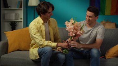 Evde homoseksüel bir çiftin fotoğrafı. Bir tanesi eşine sevgi ve sevgisini ifade etmek için çiçek ve buket verdi. Erkekler, partnerler şefkatle sarılıyorlar. LGBT, eşitlik, eğitim içeriği.