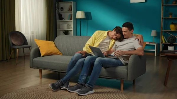 一对同性恋夫妇在家里拍的全景照片一个是坐在沙发上 在笔记本电脑上工作 向旁边的伴侣解释一些事情 他们在拥抱 Lgbt — 图库照片
