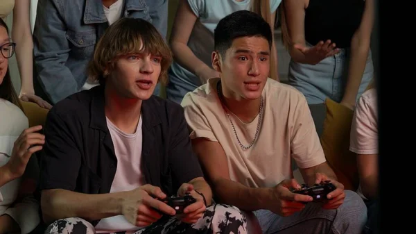 一群青少年 年轻人 朋友们围坐在电视前 站在电视前欢呼 鼓励着两个人 男孩们玩着游戏机 玩游戏 游戏者 青少年 青年内容 — 图库照片