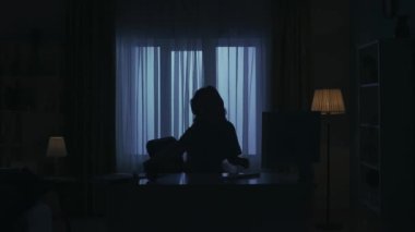 Karanlık dairedeki kadın siluetinin portresi. Günlük yaratıcı yaşam konsepti. Günlük kıyafetli bir kadın odada masa başında oturmuş bilgisayar başında çalışıyor, kağıt dökümanlarını kontrol ediyor, kahve içiyor..