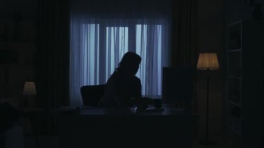 Karanlık dairedeki kadın siluetinin portresi. Günlük yaratıcı yaşam konsepti. Günlük kıyafetli bir kadın odada oturmuş bilgisayar başında yorgun ve sinirli bir şekilde kağıtları masaya fırlatıyor..