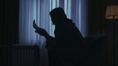 Karanlık dairedeki kadın siluetinin portresi. Günlük yaratıcı yaşam konsepti. Ekoseye bulanmış bir kadın ısıtıcının yanındaki sandalyeye oturur, üşüdüğünü hisseder, ellerini ısıtır, talimatları okur.