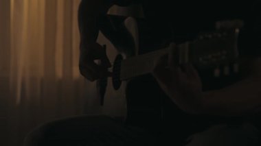 Oturma odasında oturan ve akustik gitarla şarkı çalan bir adamın yakın çekimi. Hobi zamanının tadını çıkarıyor. Karanlık dairedeki erkek siluetinin portresi. Günlük hayat yaratıcı konsept.