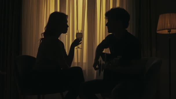 暗いアパートのシルエットの人の肖像画 日常生活のクリエイティブコンセプト リビングルームに座っている若いカップル ギターを弾く男 女性はワインのグラスを聞き 飲みます — ストック動画