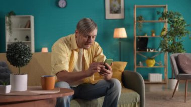 Bir adam gergin bir şekilde bir video izliyor, bir spor programı, çevrimiçi bir oyun oynuyor, zafer dansı yapıyor, galibiyeti kutluyor. Sıradan kıyafetler giymiş, elinde telefonla evdeki kanepede oturan bir adam.