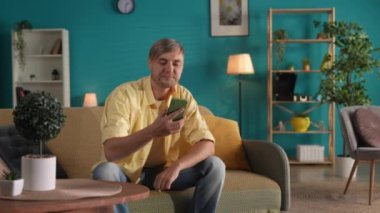 Evdeki kanepede oturan bir adam akıllı telefondaki bir video konuşmasında konuşuyor. Adam selamlaşmak için elini sallıyor, odanın içini gösteriyor ve hoşça kal diyor.