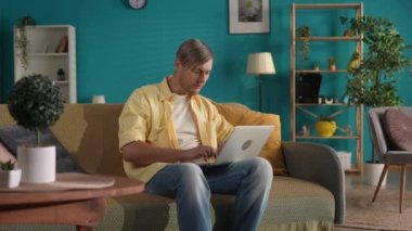 Günlük kıyafetli bir adam oturma odasındaki kanepede dizüstü bilgisayarıyla oturuyor. Bir adam çalışır, mesaj yazar, mesaj yazar, sohbet eder, online oyunlar oynar. Uzak çalışma, serbest çalışan.