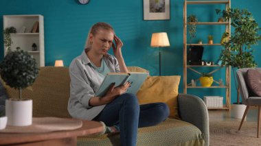 Kadın evde kanepede otururken kitap okuyor. Bir kadın okuduklarını düşünerek elini başının yanında tutuyor. Aile mutluluğu, rahatlık, evde dinlenme.