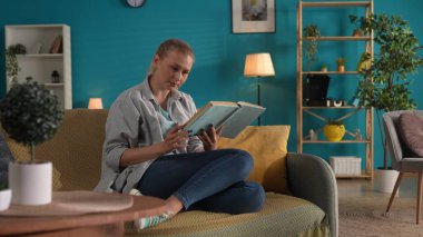 Kadın evde kanepede otururken kitap okuyor. Aile mutluluğu, rahatlık, evde dinlenme.