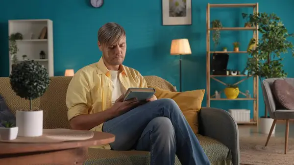 一个满脸沉思 手里拿着一本书的男人坐在客厅的沙发上 这个人在想他所读到的东西 家庭幸福 在家里休息的概念 — 图库照片