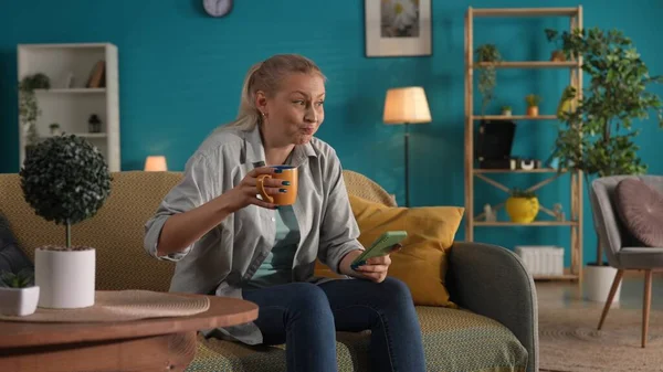 一个女人坐在沙发上的客厅里 手里拿着智能手机 一个女人在喝茶 喝杯子里的咖啡 — 图库照片